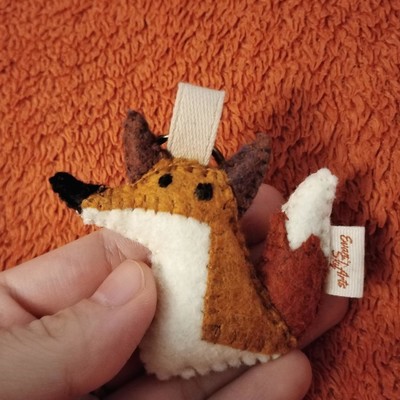 Square photo of my left hand holding a very small felt fox.
It's a keychain, and very cute.
The background is an orange blanket, kind of a similar colour to the orange part of the fox's tail.