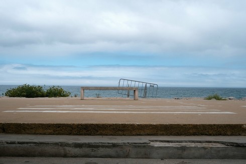 View of the beach promenade at Costa da Caparica. Up front an empty Bench and metallic barrier leaning on it. Behind the bench, the sea in an partially overcast day.